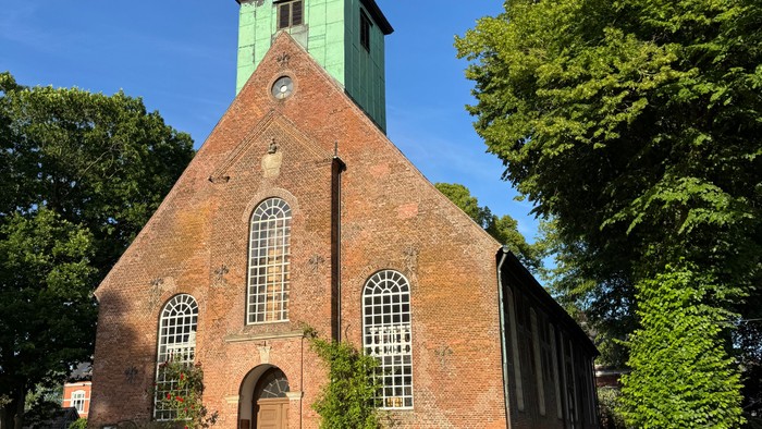 Historische Backsteinkirche mit grünem Turm, umgeben von üppigem Grün und Wildblumen unter klarem blauem Himmel.