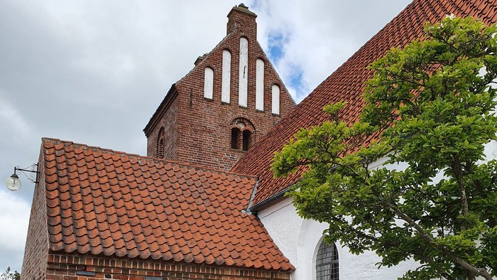 En lille kirke med røde teglsten og hvidkalket tårn med træbank og bord i forgrunden