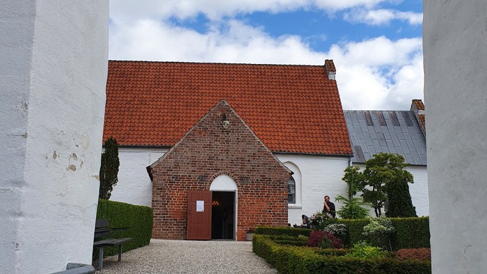 En lille rødfærgede kapel med indgang og bænke foran, omgivet af grønne buske og en sti.