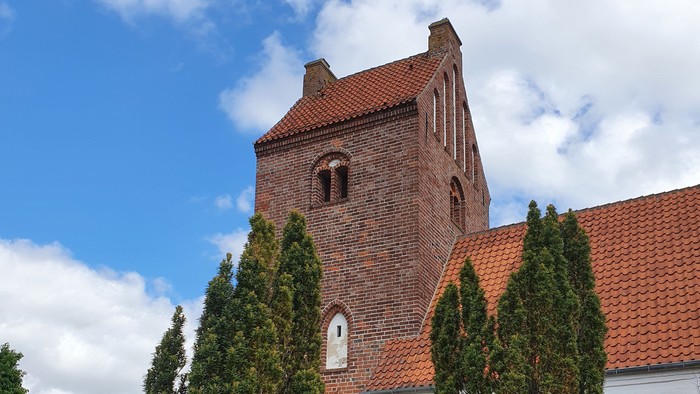 En rød murstenskirke med spiret og klokkehuset, omgivet af grønne træer og græs.