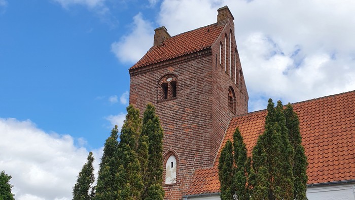 En rød murstenskirke med spiret og klokkehuset, omgivet af grønne træer og græs.