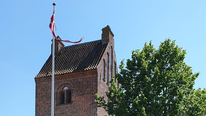 En rød murstenskirke med tårn og flagstang foran, omgivet af en hvid mur og træer
