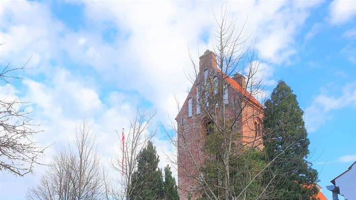 En rød murstenskirke med tårn og klokke, omgivet af træer og buske