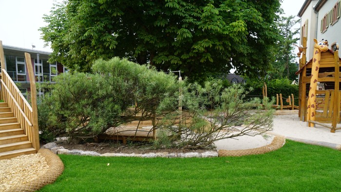 Üppiger Gartenbereich mit gepflegten Sträuchern, Kieswegen und einer hölzernen Spielstruktur neben einem Haus.