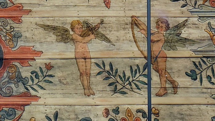 Zwei engelhafte Figuren mit Flügeln, die inmitten aufwendiger Verzierungen Musikinstrumente spielen.