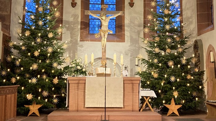 In der Kirche stehen zwei Weihnachtsbäume mit Lichtern und Sternen, davor ein Altar mit Kruzifix und Kerzen.