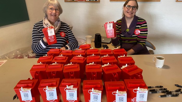 To kvinder siddende ved bord med mange røde indsamlingsbøsser fra Folkekirkens Nødhjælp foran sig
