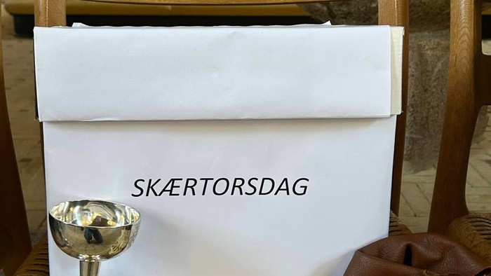 Kalk, brød og læsersæk med mønter anbragt på stol foran kasse med teksten Skærtorsdag 