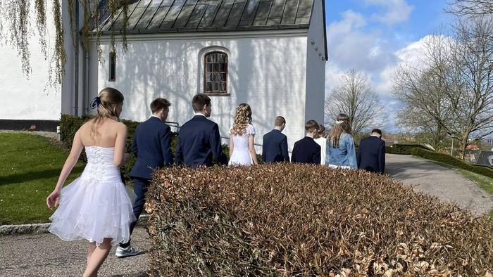 Ni unge mennesker i konfirmationstøj set fra ryggen gående langs hvid kirke
