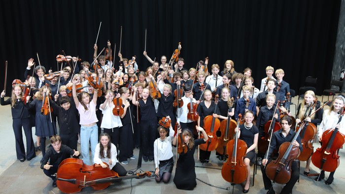 Eine vielfältige Jugendkapelle posiert gemeinsam mit ihren Streichinstrumenten auf der Bühne.