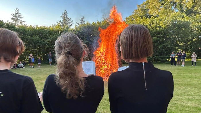 Tre sortklædte unge kvinder set fra ryggen syngende med front mod sankthansbål i stor have. Flere mennesker i baggrunden. 