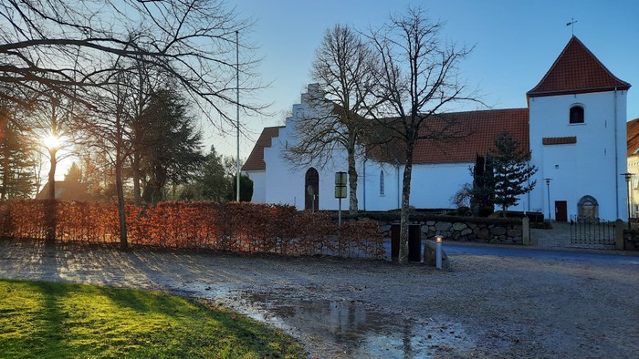 En smuk kirke med tårn og rødt tag, omgivet af træer og en murstenge hegn.