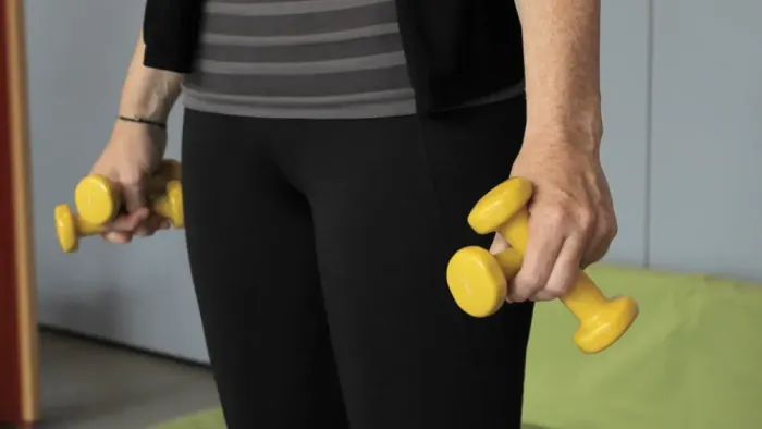 Person hält gelbe Kurzhanteln und steht auf einer grünen Matte.