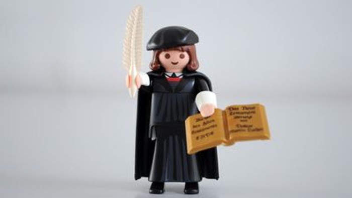 Eine kleine Figur in einer Robe hält ein Pergament und eine Feder.