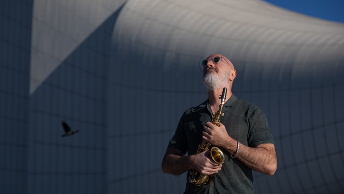 Ein bärtiger Mann spielt unter einer großen, modernen Stoffkonstruktion im Freien Saxophon, während ein Vogel in der Nähe fliegt.