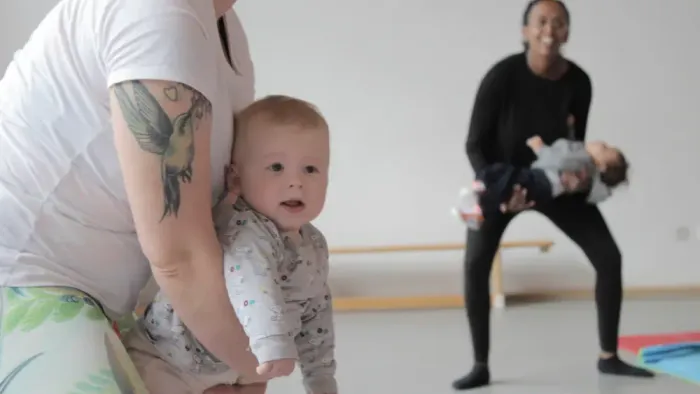 Eine Frau hält ein Baby, während eine andere Frau im Hintergrund Sport macht.
