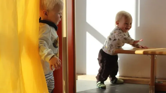 Zwei kleine Kinder, die drinnen spielen – eines steht neben einem gelben Vorhang, das andere in der Nähe eines hölzernen Tisches.