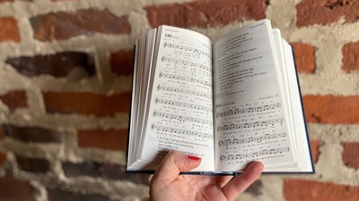 En hånd holder en åben noterbog med musiknoter foran en mur af røde teglsten.