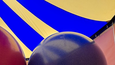 Zwei große Ballons auf dem Rasen, einer rot und einer blau, mit einem bunten Zelt im Hintergrund.