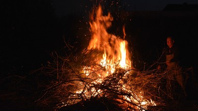 Ein Lagerfeuer brennt in der Dunkelheit, eine Person sitzt daneben.