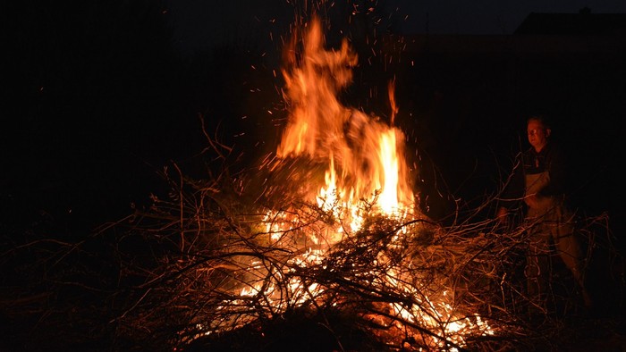 Ein Lagerfeuer brennt in der Dunkelheit, eine Person sitzt daneben.