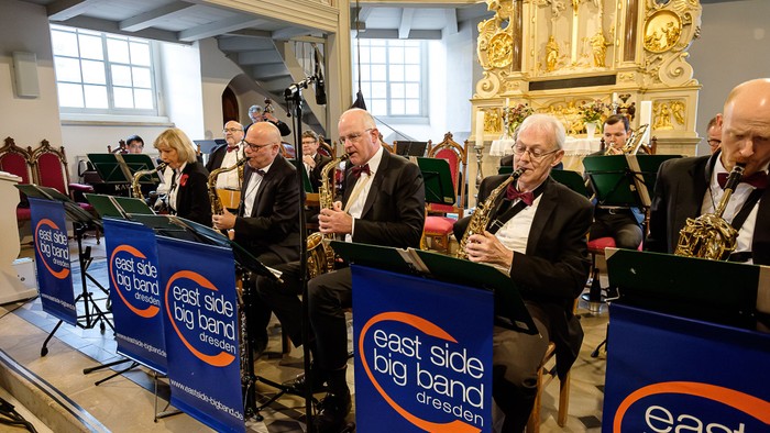 Eine Gruppe von Musiker:innen der East Side Big Band Dresden spielt Blasinstrumente auf der Bühne in einem Innenraum.