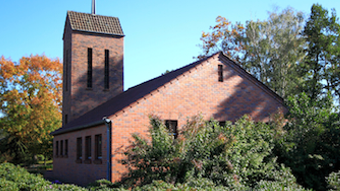Das Bild zeigt eine rote Ziegelkirche mit hohem Turm und Kreuz, umgeben von Bäumen und Büschen.
