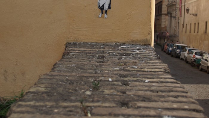 Ein Bild einer Person an einer gelben Wand über Kopfsteinpflaster.