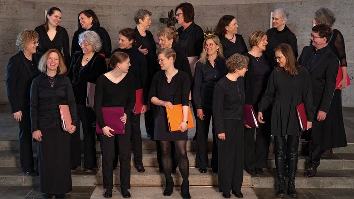 Eine professionelle Chorgruppe steht in formeller Kleidung zusammen, hält Notenblätter und Taktstöcke.