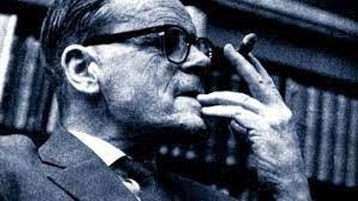 En ældre mand med briller og skæg, iført jakkesæt, holder en pibe i hånden.