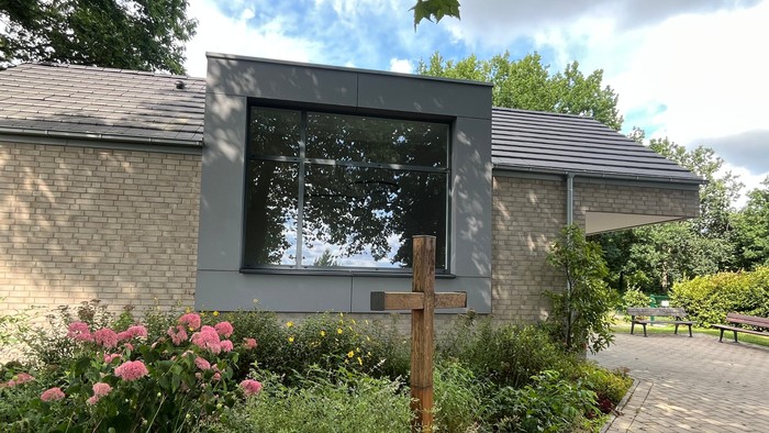 Hier sieht man eine Seitenansicht der Laggenbecker Friedhofskapelle mit dem großen Fenster und im Pflanzbeet davor steht das Holzkreuz der alten Kapelle.