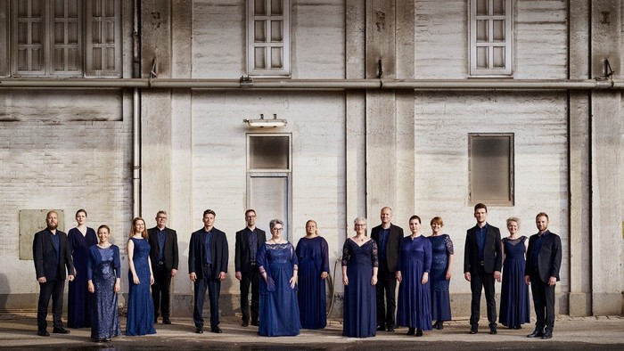 En gruppe af mænd og kvinder i formelle blå tøjer står foran et stort, koldt og gråt stenhus med flere vinduer.