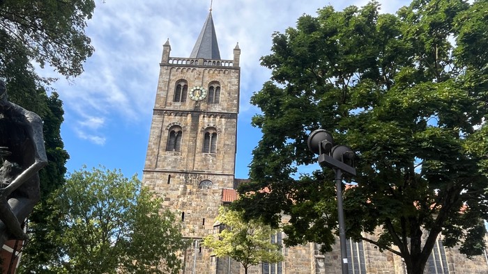 Das Bild zeigt links den großen Glockenturm aus Sandstein und nach rechts das weitere Kirchengebäude mit hohe Fenstern, teilweise von Bäumen im Vordergrund verdeckt.
