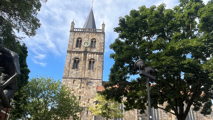Das Bild zeigt links den großen Glockenturm aus Sandstein und nach rechts das weitere Kirchengebäude mit hohe Fenstern, teilweise von Bäumen im Vordergrund verdeckt.