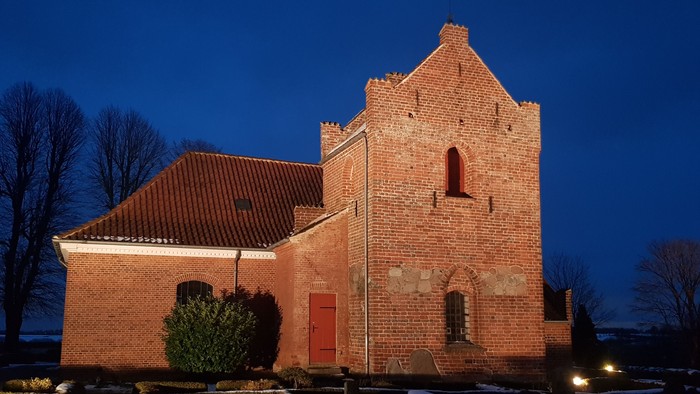 En rød murstenskirke med spids tak og en lille tårn i mørket.