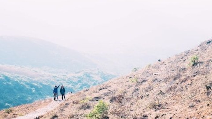 Zwei Wanderer steigen einen nebligen, kargen Hang hinauf, während in der dunstigen Ferne Berge zu sehen sind.