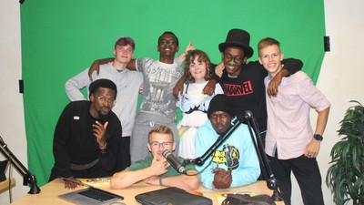 Eine Gruppe von Menschen posiert vor einem grünen Hintergrund, einige halten Mikrofone und Laptops