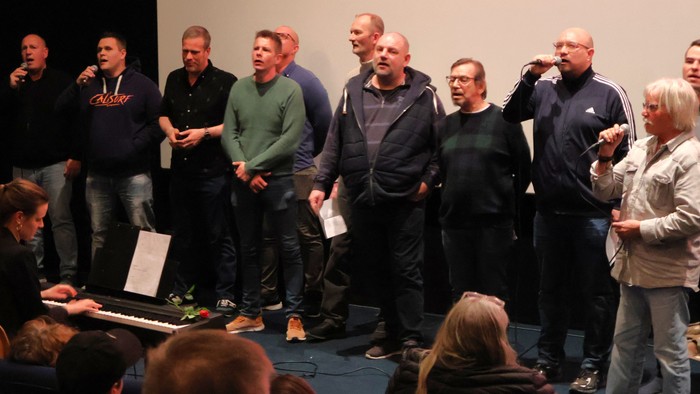 En gruppe mænd og en kvinde, der synger eller optræder sammen med en pianist i en publikumssituation.