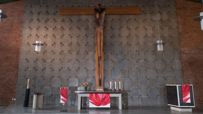 Die Kirche zeigt einen Altar mit Kreuz und Kerzen, flankiert von zwei roten Teppichen und einem Bildschirm.