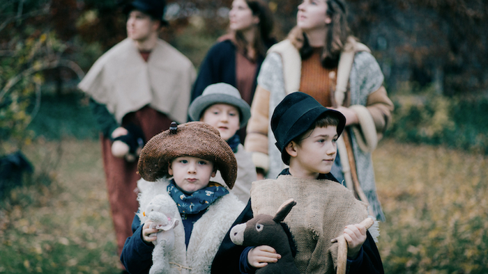 Kinder in historischer Kleidung, eines mit einem Spielzeug in der Hand, gehen mit Erwachsenen über eine grasbewachsene Fläche.