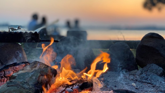 En hyggelig strandbål ved kysten med flammende ild mellem stenene under solnedgang, omgivet af mennesker, der slapper af.