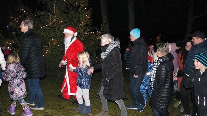 En gruppe børn og voksne samles om julemanden i en park med julelys.