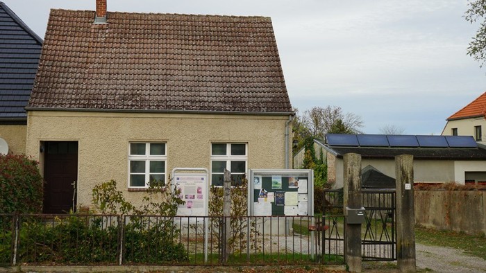 Ein kleines Haus mit braunem Ziegeldach, davor ein Zaun und zwei Infotafeln.