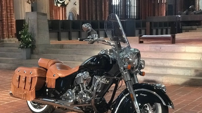 Ein schlankes schwarzes Cruiser-Motorrad mit hellbraunen Ledertaschen, das drinnen in der Nähe von Treppen und hölzernen Kirchenbänken geparkt ist.