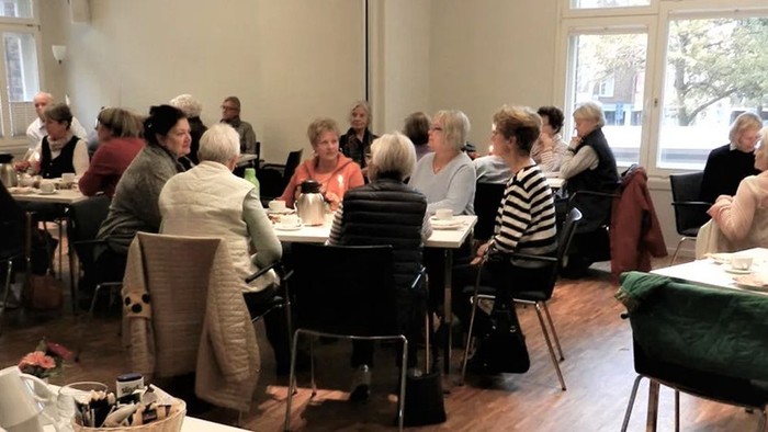 Eine Gruppe älterer Menschen sitzt an Tischen und isst gemeinsam in einem hellen Raum.