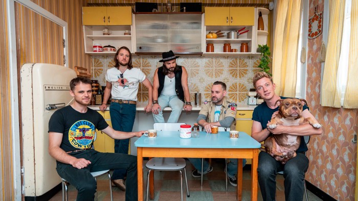 En gruppe på seks personer og en hund samles i et retro-inspireret køkken.