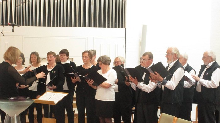Et kor optræder i en kirke, hvor medlemmerne holder sangbøger, og en dirigent leder dem.