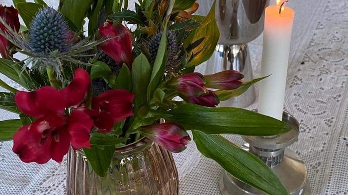 En blomsterstang i glasvas og en lystet lysestage på bord med hvid spidsbinde.