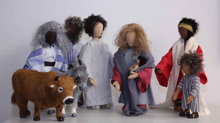 Eine Gruppe von Puppen und ein Kuhfigürchen, alle in verschiedene Gewänder gekleidet, stehen vor einer weißen Wand.