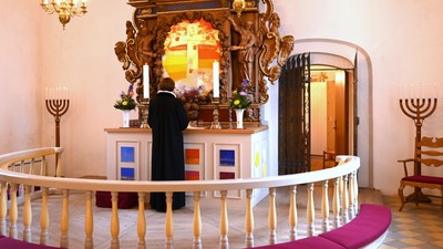 En mand i sort kåbe står ved et prægtigt alter med glasmosaik og krystallysekroner i en kirke.